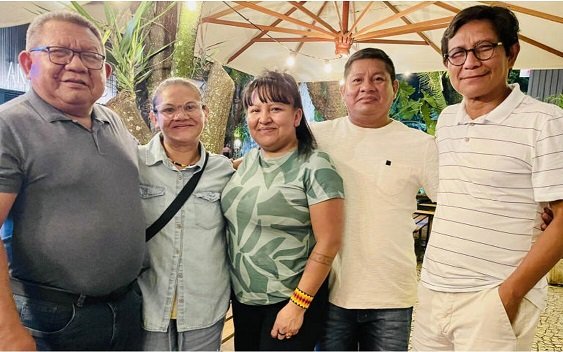Cinco pessoas posam juntas, sorrindo, em um ambiente ao ar livre com plantas e uma cobertura de madeira ao fundo. O grupo é composto por duas mulheres e três homens